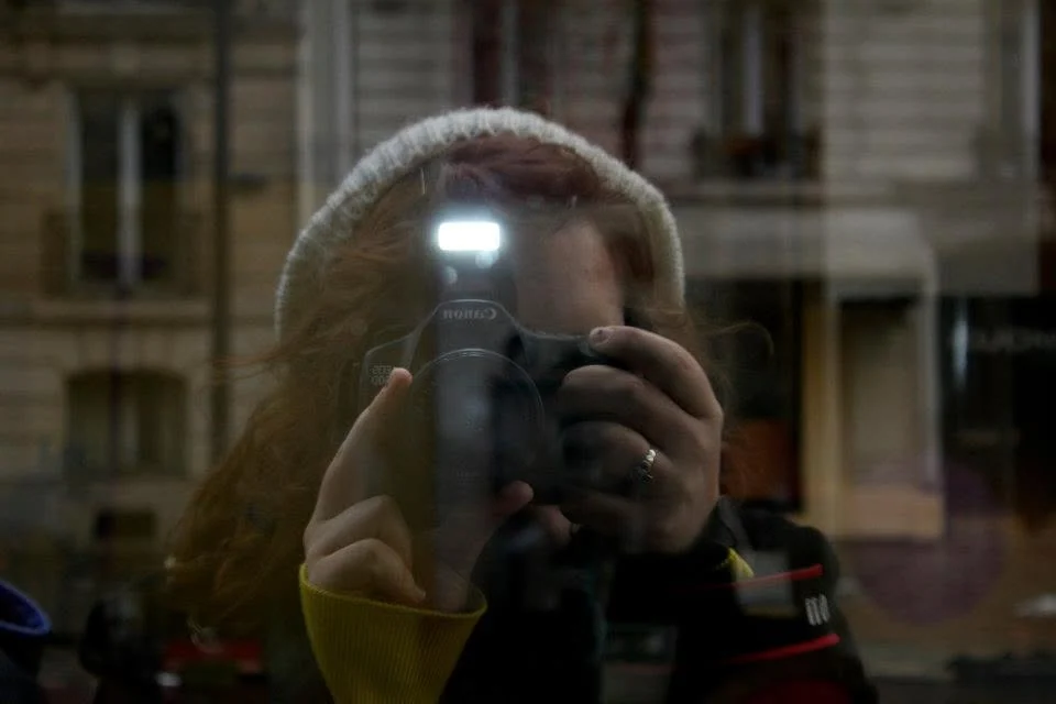 Alex photographing her own reflection in a Paris shop window in 2013, in a knit hat, Canon held up to her face